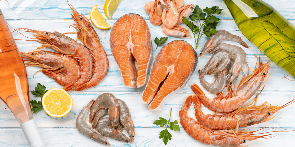 Fresh seafood assortment with salmon fillets, whole prawns, raw shrimp, lemon slices, parsley, and a bottle of oil arranged on a light wooden surface.