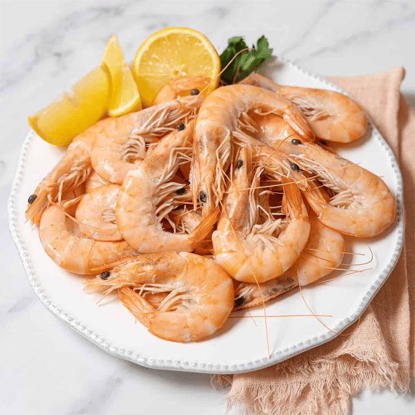 A plate of cooked shrimp garnished with lemon wedges and parsley, served on a white dish with a light cloth on a marble surface.