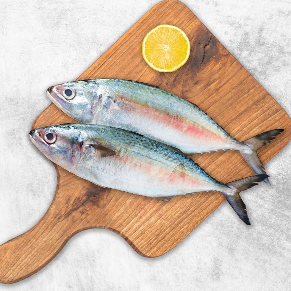 Two fresh whole fish placed on a wooden cutting board with a slice of lemon on top, set against a light background